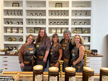 Load image into Gallery viewer, Five women in aprons standing in front of a shelf with jars and bottles in a store setting.